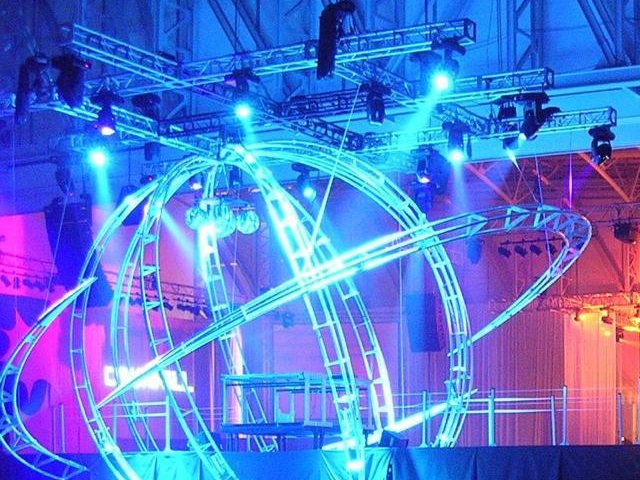 Photo of the built truss sphere at the event: blue-lit curved orbit truss arcs beside the stage in a dark hall