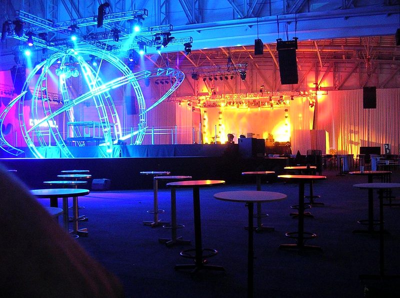 Blue-lit truss sculpture and stage lighting rig in an event hall set with cocktail tables